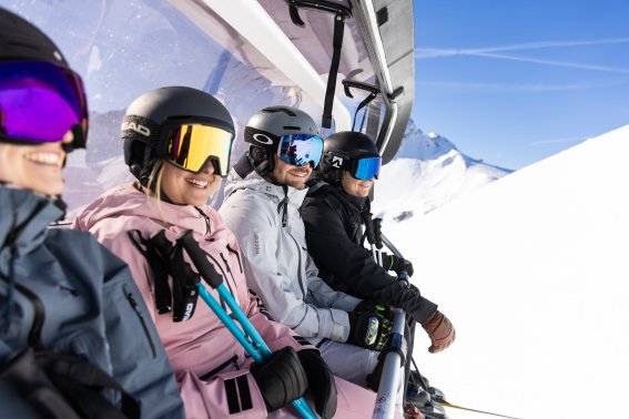 Vier Freunde lächeln gemeinsam während der Sesselliftfahrt im sonnigen Skigebiet rund um Oberstdorf. Vier Freunde sitzen gut gelaunt im Sessellift und genießen die gemeinsame Auszeit zwischen ihren Abfahrten.