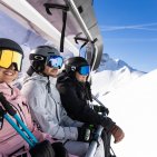 Drei Freunde genießen die Sesselliftfahrt mit weitem Bergblick im sonnigen Skigebiet bei Oberstdorf. Drei Skifahrer fahren gemeinsam mit dem Sessellift bergauf und blicken voller Vorfreude auf den nächsten Run.