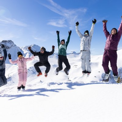 Sechs Freunde springen gemeinsam in die Luft und feiern ihren Wintertag vor der eindrucksvollen Bergkulisse.