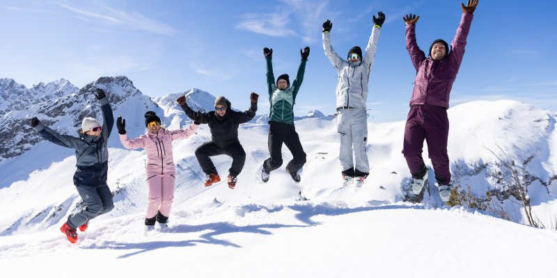 Sechs Freunde springen gemeinsam in die Luft und feiern ihren Wintertag vor der eindrucksvollen Bergkulisse.