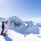 Zwei Skifahrerinnen stehen am Rand der Piste und genießen das sonnige Winterpanorama bei Oberstdorf. Ein gemeinsamer Stopp mit Wow-View: Vor den beiden Skifahrerinnen öffnet sich die ganze Weite der Bergwelt.