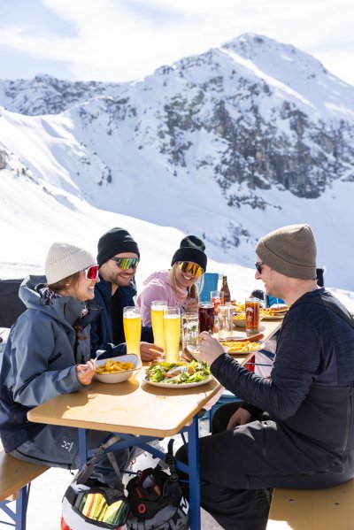Vier Freunde sitzen beim Mittagessen auf einer sonnigen Bergterrasse vor verschneiter Gipfelkulisse. Vier Freunde genießen ihre gemeinsame Einkehr auf der Terrasse – mit leckerem Essen und bestem Bergblick.