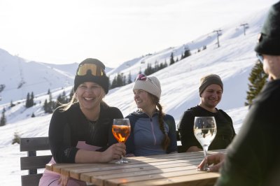 Die Freundesgruppe verbringt einen entspannten Moment mit Getränken, Wintersonne und Blick auf die verschneiten Pisten.