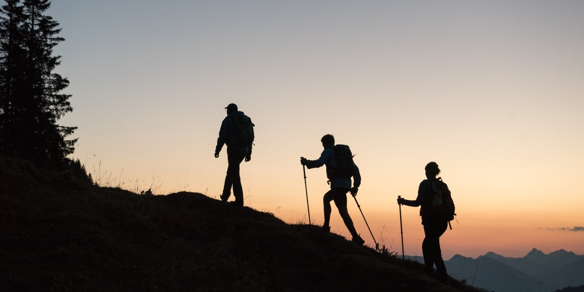 Drei Wanderer sind bei Sonnenaufgang als Silhouetten auf einem Berggrat zu sehen.