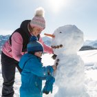Eltern und Kind genießen den Schnee in den Allgäuer Hörnerdörfern. Ein Elternteil trägt ein Kind durch die verschneite Berglandschaft. Die spielerische Szene steht für gemeinsame Wintermomente in den Hörnerdörfern.