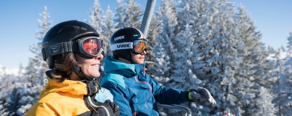 Skifahrer fahren mit dem Sessellift bergauf und genießen den Blick auf die verschneite Landschaft. Das Motiv zeigt einen entspannten Moment zwischen den Abfahrten.