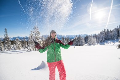 Frau genießt den Wintertraum im Schnee der Allgäuer Hörnerdörfer. Eine Urlauberin wirft begeistert Schnee in die Luft. Blauer Himmel und tief verschneite Berge schaffen einen unbeschwerten Wintermoment.
