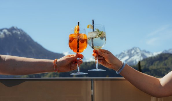 Aperitif auf der Dachterrasse Zwei Personen stoßen auf der Dachterrasse mit ihren Drinks an. Das linke Glas ist gefüllt mit einem Aperol Spritz und das rechte mit einem Hugo. Im Hintergrund sieht man das wunderschöne Bergpanorama.