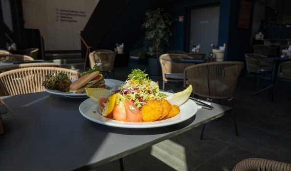 Räucherlachs mit Rösti Auf dem Tisch steht ein Teller mit frischem Räucherlachs und knusprigen Röstis.Im Hintergrund sieht man einen Teller mit einem Panini und die Gastronomie der Therme.