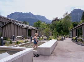 Eine Person mit blonden Haaren und einem blauen Handtuch läuft durch das Kneippbecken in der Therme. Im Hintergrund sind die Berge zu sehen, während einige grün beleuchtete Bäume für eine stimmungsvolle Atmosphäre sorgen.