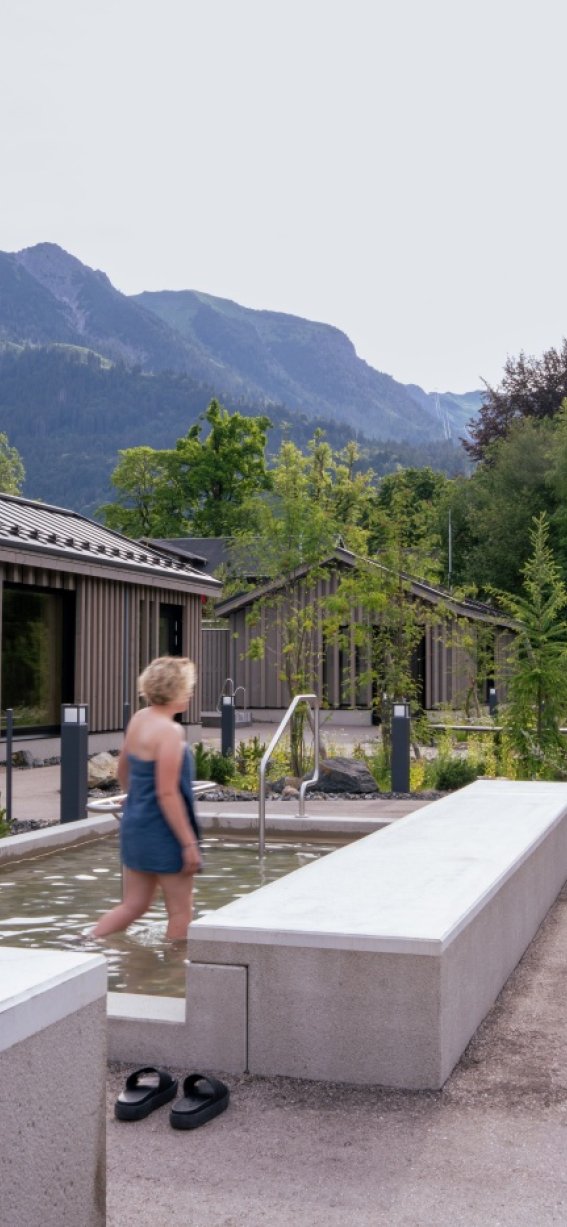 Eine Person mit blonden Haaren und einem blauen Handtuch läuft durch das Kneippbecken in der Therme. Im Hintergrund sind die Berge zu sehen, während einige grün beleuchtete Bäume für eine stimmungsvolle Atmosphäre sorgen.