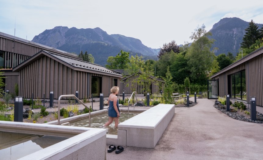 Kneippbecken Therme Eine Person mit blonden Haaren und einem blauen Handtuch läuft durch das Kneippbecken in der Therme. Im Hintergrund sind die Berge zu sehen, während einige grün beleuchtete Bäume für eine stimmungsvolle Atmosphäre sorgen.