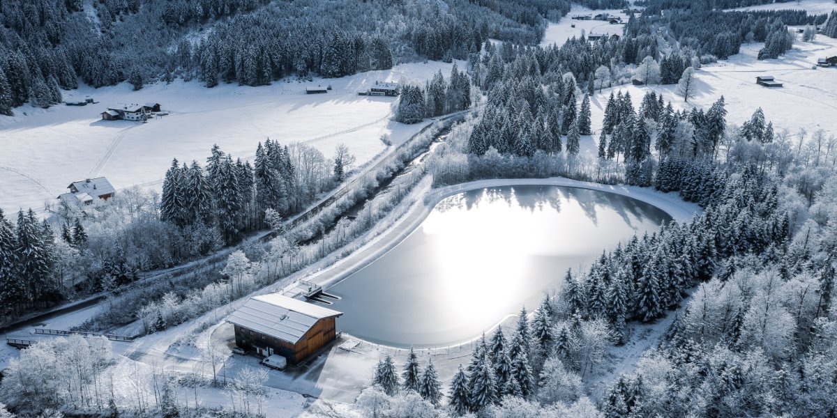 Eine malerische Winterlandschaft zeigt einen gefrorenen Teich, umgeben von schneebedeckten Bäumen und Hütten in der Ferne.