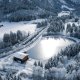 Eine malerische Winterlandschaft zeigt einen gefrorenen Teich, umgeben von schneebedeckten Bäumen und Hütten in der Ferne.