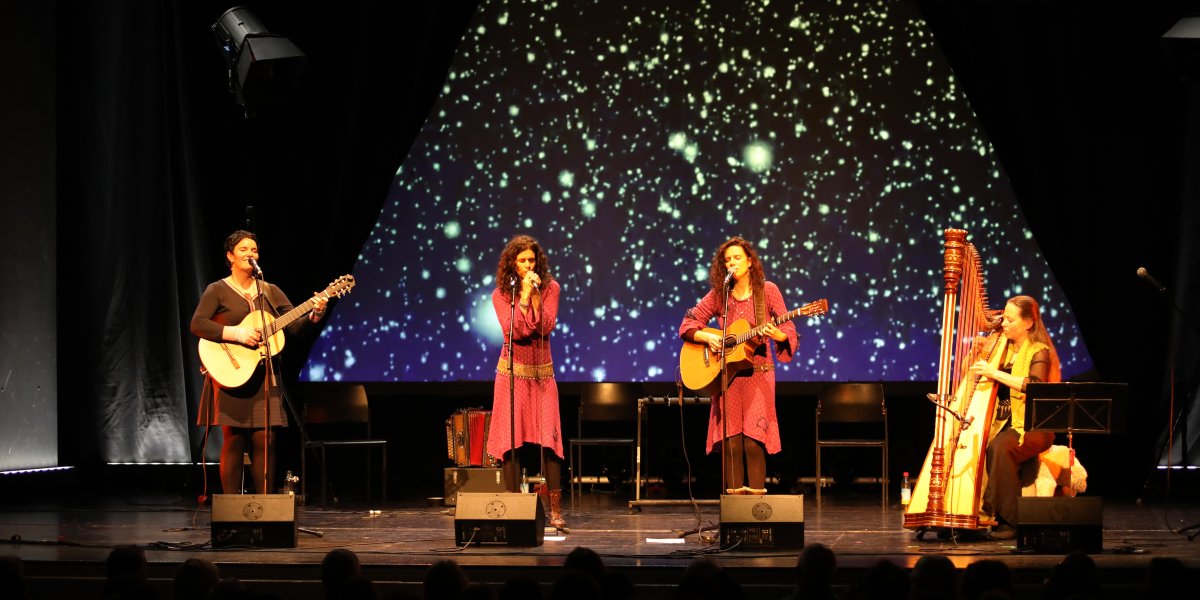 Musikerinnen performen gemeinsam auf der Bühne mit Instrumenten und beeindruckendem Lichtspiel im Hintergrund.