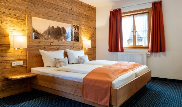 Das Zimmer zeigt ein Bett mit zwei Kissen. Neben dem Bett steht ein Tisch. Tageslicht kommt durch das Fenster.