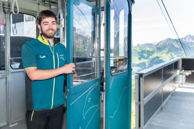 Kabinenbegleiter Ein Mitarbeiter öffnet die Tür einer Seilbahn auf einem Berggipfel. Die Landschaft ist grün und hügelig.