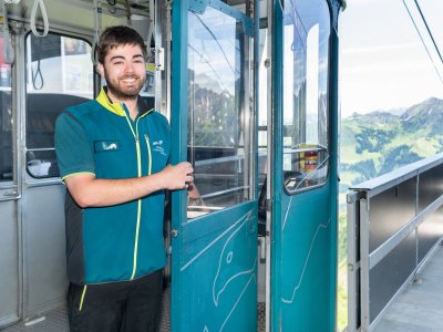 Ein Mitarbeiter öffnet die Tür einer Seilbahn auf einem Berggipfel. Die Landschaft ist grün und hügelig.