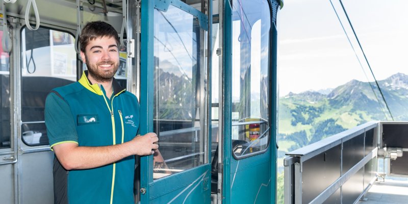 Ein Mitarbeiter öffnet die Tür einer Seilbahn auf einem Berggipfel. Die Landschaft ist grün und hügelig.