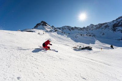 Ein Skifahrer im roten Anzug fährt die verschneite Piste hinunter. Die Sonne strahlt am blauen Himmel.