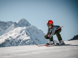 Kleine Rennsemmel Ein Kind fährt auf Ski den Hang hinunter, umgeben von schneebedeckten Bergen an einem sonnigen Tag.