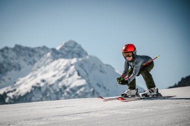 Ein Kind fährt auf Ski den Hang hinunter, umgeben von schneebedeckten Bergen an einem sonnigen Tag.