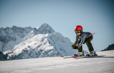 Ein Kind fährt auf Ski den Hang hinunter, umgeben von schneebedeckten Bergen an einem sonnigen Tag.