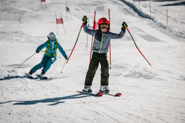 Zwei Kinder fahren Ski auf einer Schneepiste. Eines lächelt und macht eine Pose, während das andere hinterherfährt.