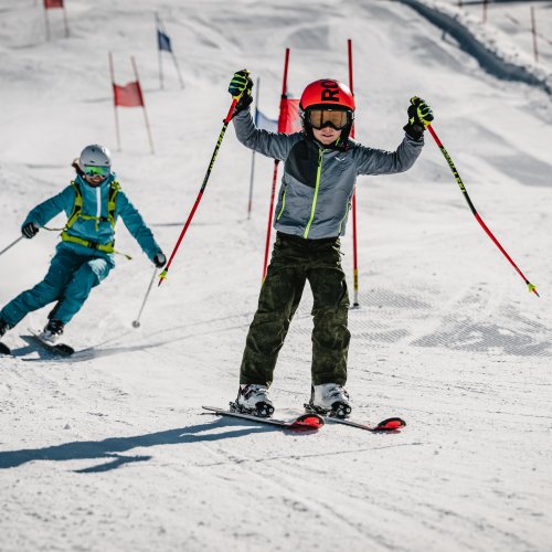 Als erster im Ziel Zwei Kinder fahren Ski auf einer Schneepiste. Eines lächelt und macht eine Pose, während das andere hinterherfährt.