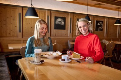 Zwei Frauen sitzen in einem Café und essen Kuchen. Sie lachen und haben Spaß in einer gemütlichen Atmosphäre.