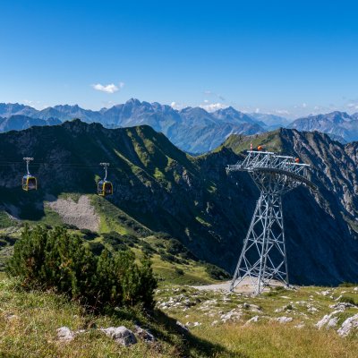 Eine Seilbahn fährt über die Alpenlandschaft an einem sonnigen Tag. Weite Aussicht auf Berge und grüne Wiesen.
