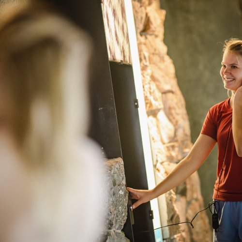 Naturkunde in der Bergschau Eine junge Frau in einem Zimmer lächelt und spricht am Telefon. Es ist eine entspannte Atmosphäre mit Steinmauern.