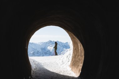 Ein Wanderer steht in einem Tunnel und blickt auf eine verschneite Berglandschaft unter klarem Himmel.