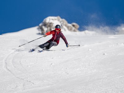 Eine Person fährt mit Schwung die Piste hinunter. Der Himmel ist blau und die Berge sind mit Schnee bedeckt.