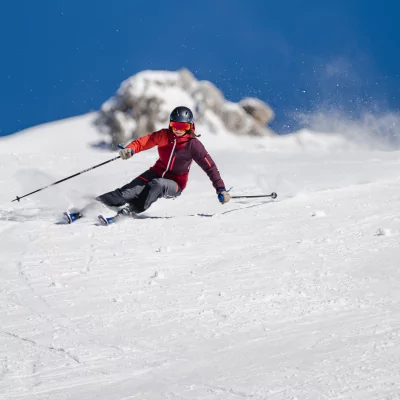 Eine Person fährt mit Schwung die Piste hinunter. Der Himmel ist blau und die Berge sind mit Schnee bedeckt.
