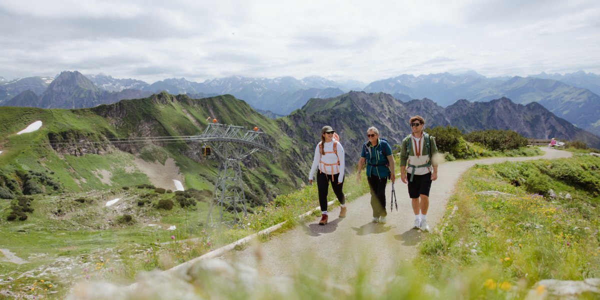 Drei Wanderer gehen einen Pfad in den Bergen und bewundern die Landschaft unter einem bewölkten Himmel.