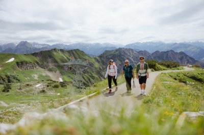 Drei Wanderer gehen einen Pfad in den Bergen und bewundern die Landschaft unter einem bewölkten Himmel.