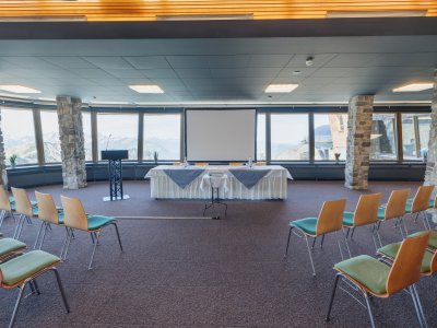 Der Raum zeigt eine Vorbereitung für eine Konferenz mit Tischen, Stühlen und einer großen Leinwand.