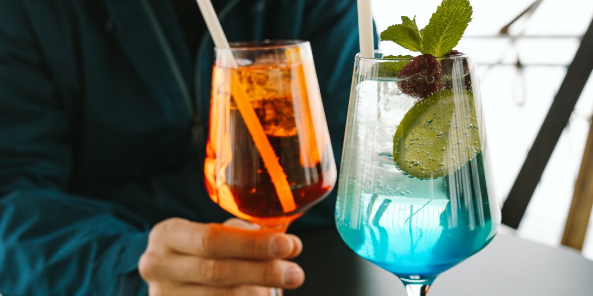 Zwei Freunde sitzen am Tisch und stoßen mit bunten Cocktails an. Der Moment ist entspannt und fröhlich.