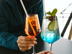 Zwei Freunde sitzen am Tisch und stoßen mit bunten Cocktails an. Der Moment ist entspannt und fröhlich.