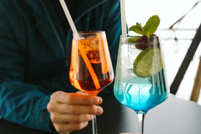 Zwei Freunde sitzen am Tisch und stoßen mit bunten Cocktails an. Der Moment ist entspannt und fröhlich.