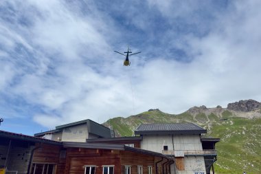 Anlieferung PV Höfatsblick Ein Hubschrauber fliegt über ein Bergdorf und transportiert Materialien für Bauarbeiten. Der Himmel ist bewölkt.