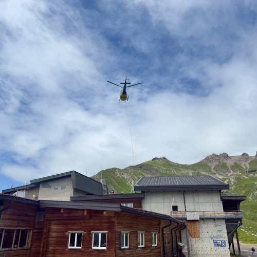 Ein Hubschrauber fliegt über ein Bergdorf und transportiert Materialien für Bauarbeiten. Der Himmel ist bewölkt.