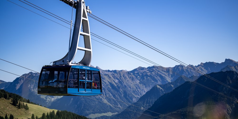 Eine Seilbahn schwebt über die Alpenlandschaft bei klarem Wetter, umgeben von hohen Bergen und grünen Wiesen. neue Kabine FBO