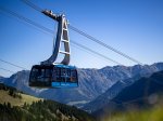 Eine Seilbahn schwebt über die Alpenlandschaft bei klarem Wetter, umgeben von hohen Bergen und grünen Wiesen.