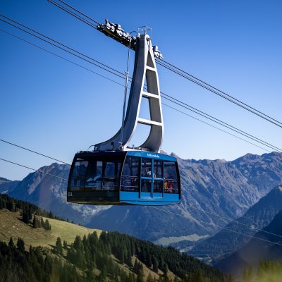 Eine Seilbahn schwebt über die Alpenlandschaft bei klarem Wetter, umgeben von hohen Bergen und grünen Wiesen.