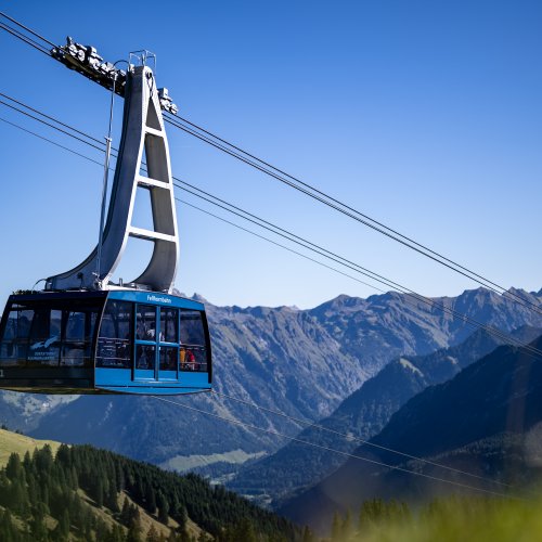 Eine Seilbahn schwebt über die Alpenlandschaft bei klarem Wetter, umgeben von hohen Bergen und grünen Wiesen.