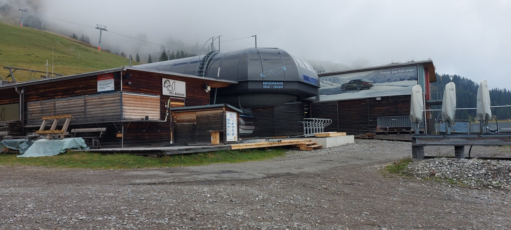 Möserbahn Sep'25 Ein modernes Gebäude in den Alpen, umgeben von Nebel, mit einem leeren Parkplatz und Holzgestellen.