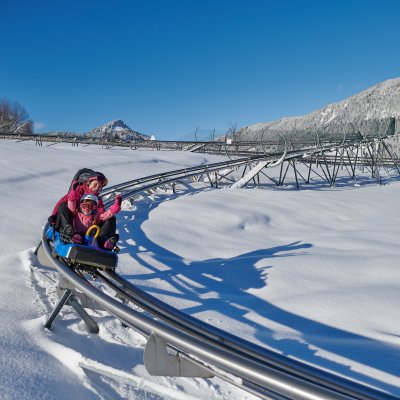 Kinder fahren mit einer Rodelbahn über schneebedeckte Landschaften. Der Himmel ist blau und die Bergkulisse beeindruckend.