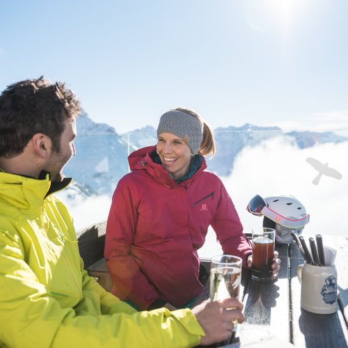 Zwei Personen in Skikleidung sitzen an einem Tisch und genießen Getränke in den Bergen an einem sonnigen Tag.